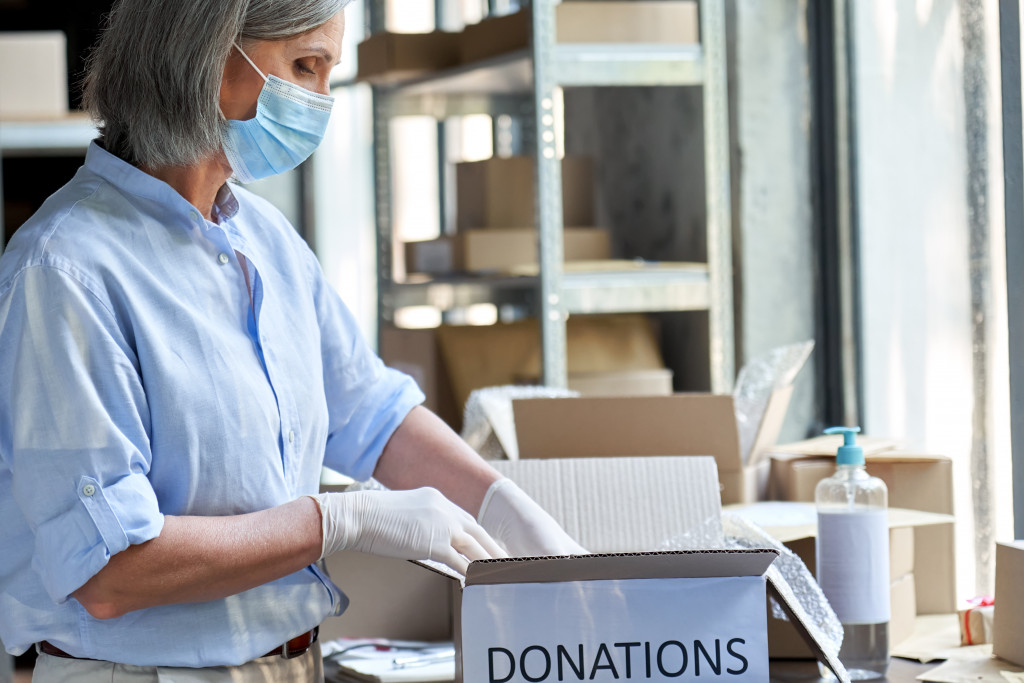 Mature female warehouse worker volunteer wearing face mask working in shipping delivery charitable stock organization packing donations box.